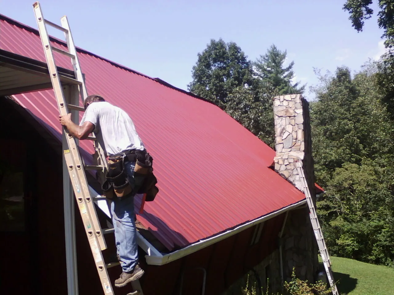 Roofer on a ladder beside a red metal roof and stone chimney — Soffit & Fascia Repair by Roofing Brothers in Wheelwright